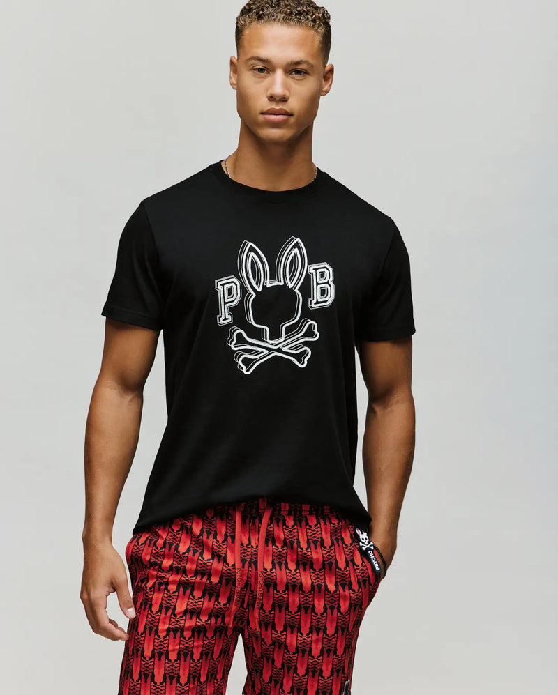 A man in a Psycho Bunny MENS BALES GRAPHIC TEE (MT0200035) with a black Big Bunny skull and crossbones design and "PB" letters, paired with red patterned shorts, stands against a plain light background.