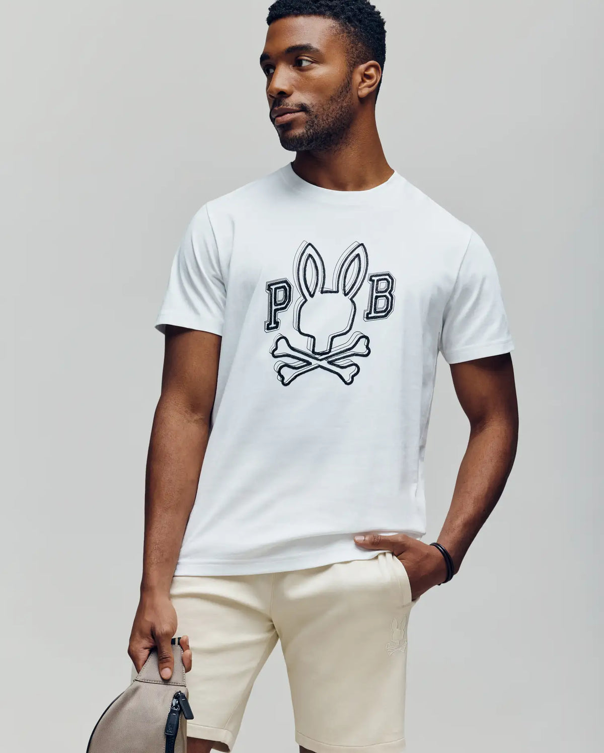 A man wears the Psycho Bunny MENS BALES GRAPHIC TEE (MT0200035), featuring a white Pima cotton fabric with a Bunny graphic and "PB" text. He pairs it with beige shorts and holds a light gray bag, styled for year-round wear.