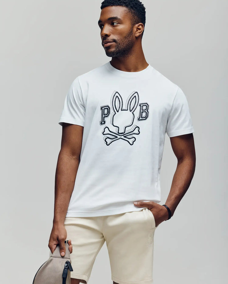 A man wears the Psycho Bunny MENS BALES GRAPHIC TEE (MT0200035), featuring a white Pima cotton fabric with a Bunny graphic and "PB" text. He pairs it with beige shorts and holds a light gray bag, styled for year-round wear.