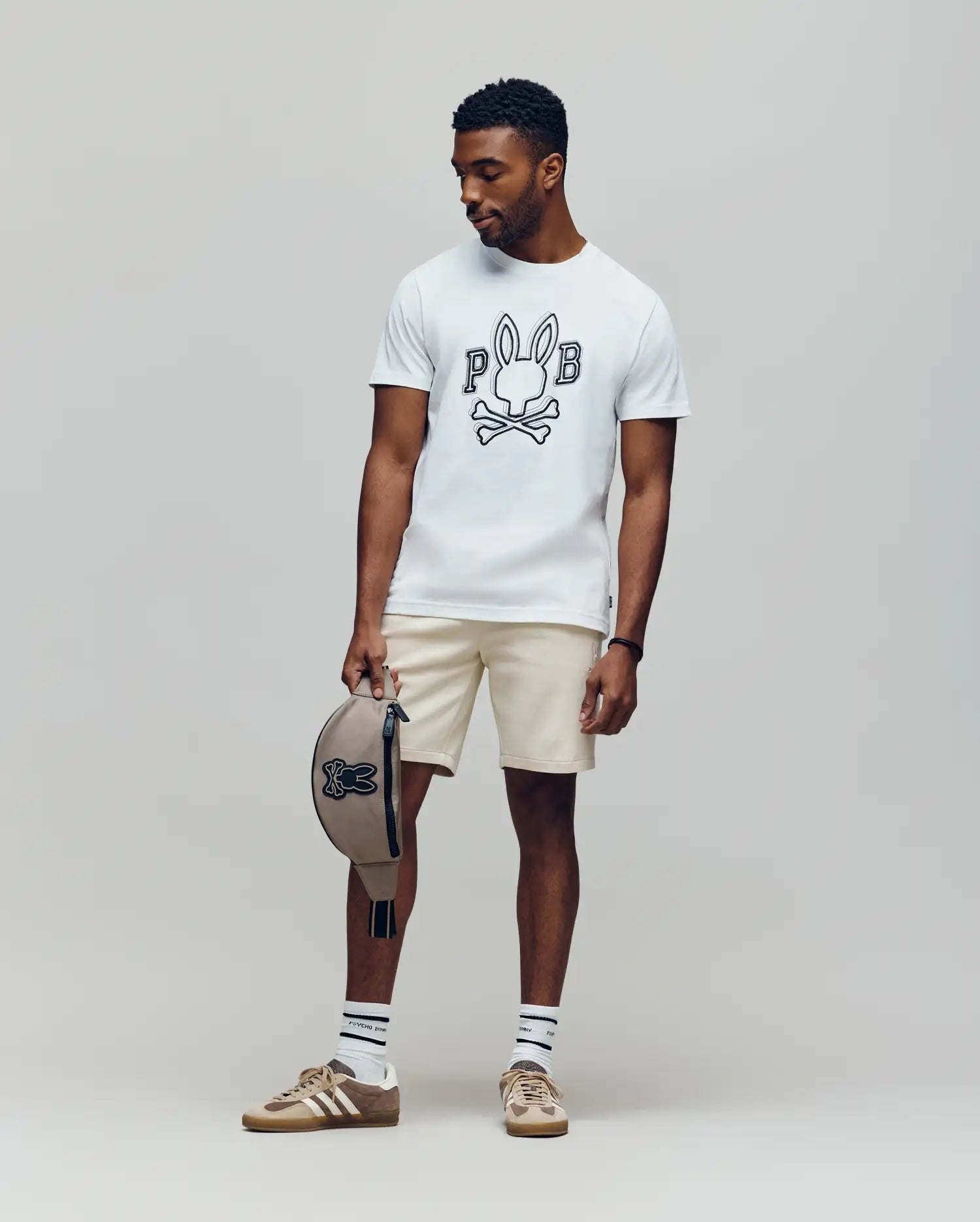 A man stands against a plain backdrop wearing Psycho Bunny’s MENS BALES GRAPHIC TEE (MT0200035) in white with a Bunny print, beige shorts, striped socks, brown sneakers, and holding a matching beige cap—ideal for year-round style.