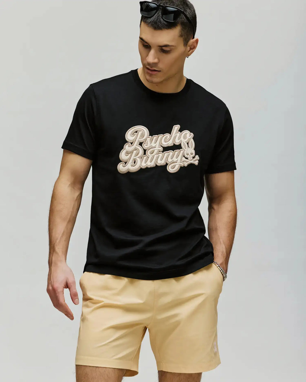 A man stands against a plain background wearing sunglasses, the Psycho Bunny MENS ELIAS GRAPHIC TEE (MT0200047) with a retro Bunny graphic and beige lettering, paired with light beige shorts, looking down with his left hand in his pocket.