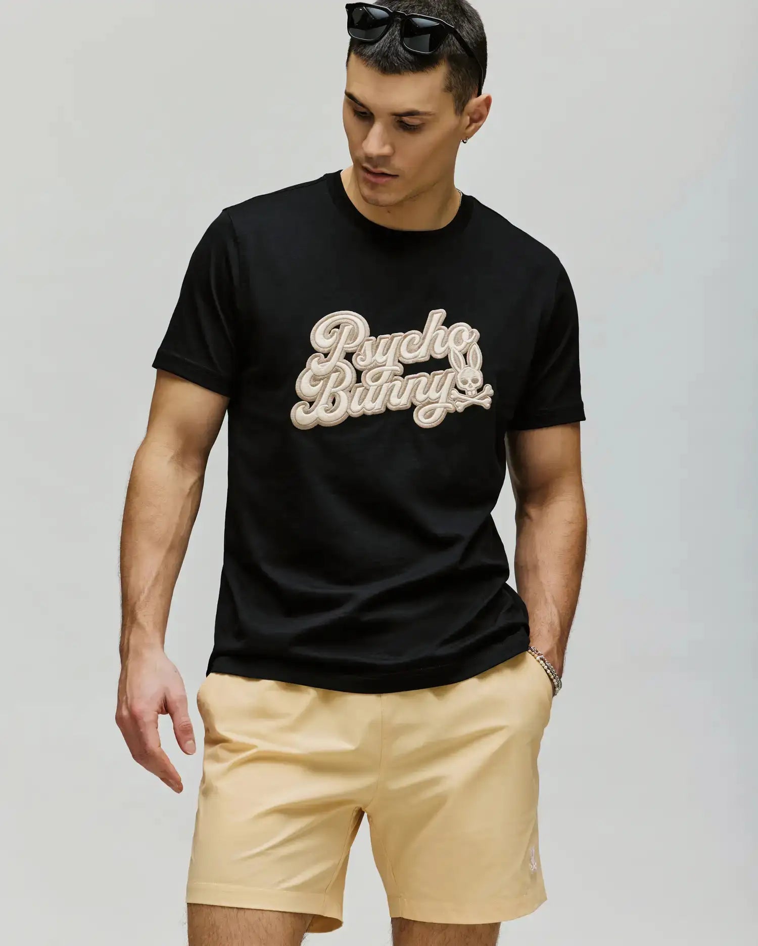 A man stands against a plain background wearing sunglasses, the Psycho Bunny MENS ELIAS GRAPHIC TEE (MT0200047) with a retro Bunny graphic and beige lettering, paired with light beige shorts, looking down with his left hand in his pocket.