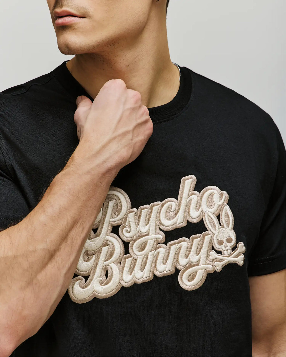 A man wears the Psycho Bunny MENS ELIAS GRAPHIC TEE (MT0200047), a black Pima cotton shirt with bold textured beige "Psycho Bunny" lettering and a retro bunny skull logo, partially covered by his raised left fist near his neck.