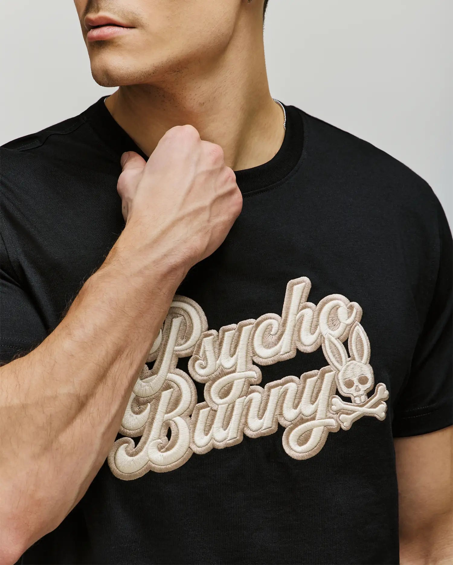 A man wears the Psycho Bunny MENS ELIAS GRAPHIC TEE (MT0200047), a black Pima cotton shirt with bold textured beige "Psycho Bunny" lettering and a retro bunny skull logo, partially covered by his raised left fist near his neck.