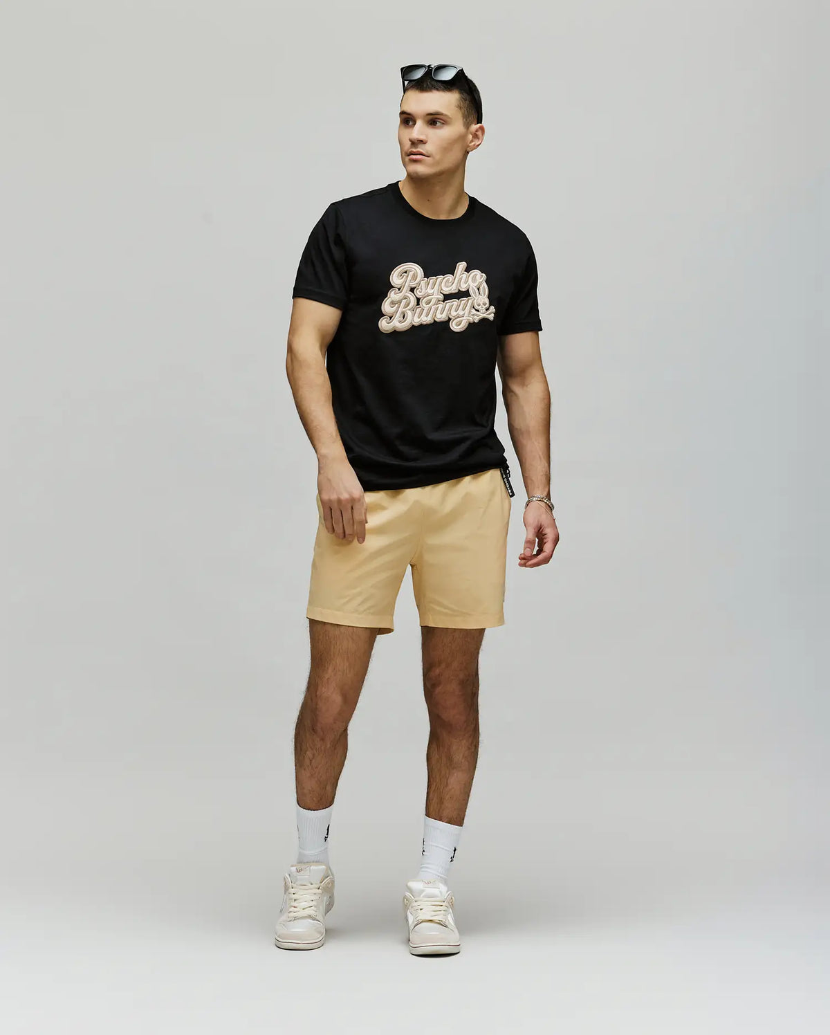 A man stands against a plain background wearing the Psycho Bunny MENS ELIAS GRAPHIC TEE (MT0200047) in black, paired with beige shorts, white sneakers and socks, and sunglasses on his head. He faces slightly to the side with one hand by his hip.