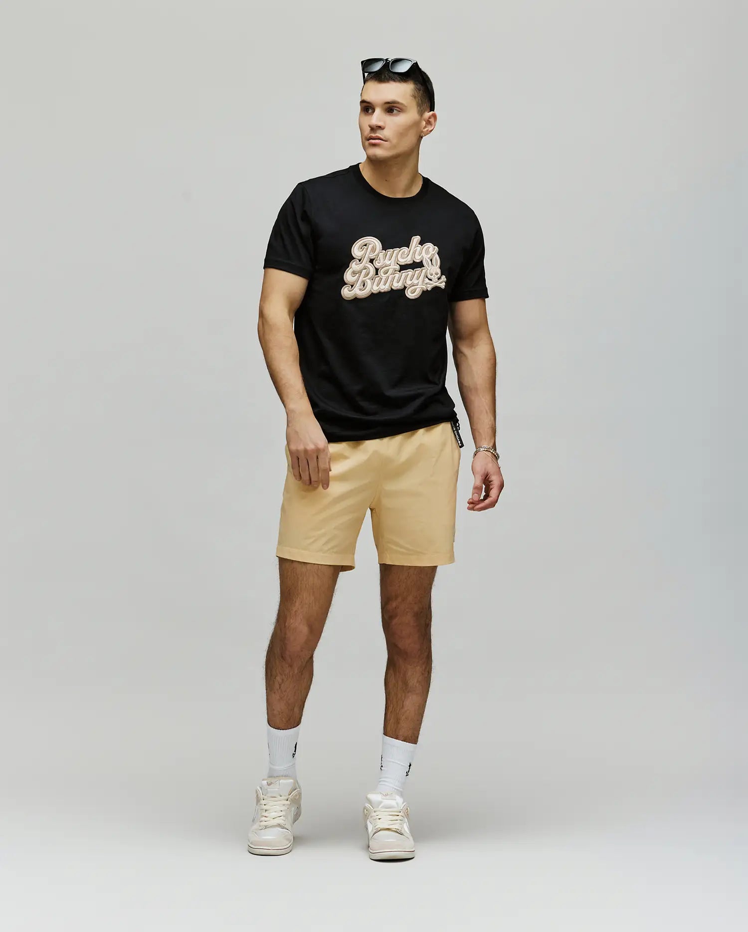A man stands against a plain background wearing the Psycho Bunny MENS ELIAS GRAPHIC TEE (MT0200047) in black, paired with beige shorts, white sneakers and socks, and sunglasses on his head. He faces slightly to the side with one hand by his hip.