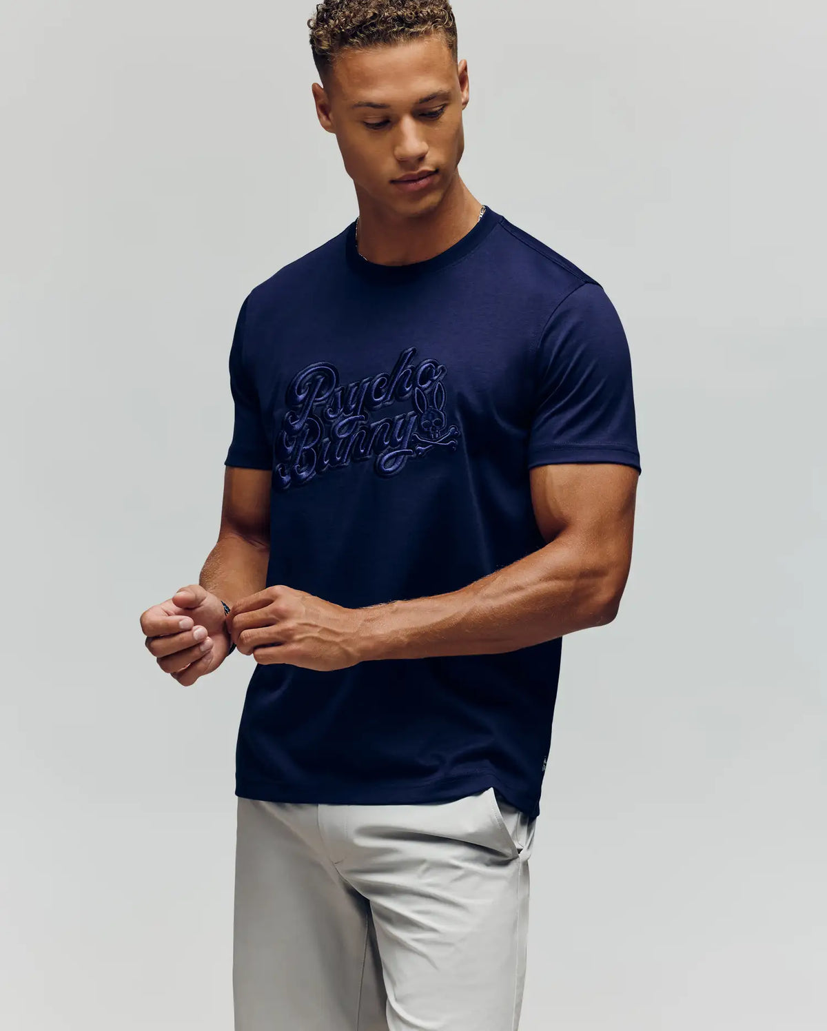 A man in light pants wears the MENS ELIAS GRAPHIC TEE (MT0200047) by Psycho Bunny—navy with embroidered script and a subtle Bunny graphic—standing against a plain light background, looking down with hands loosely clasped.