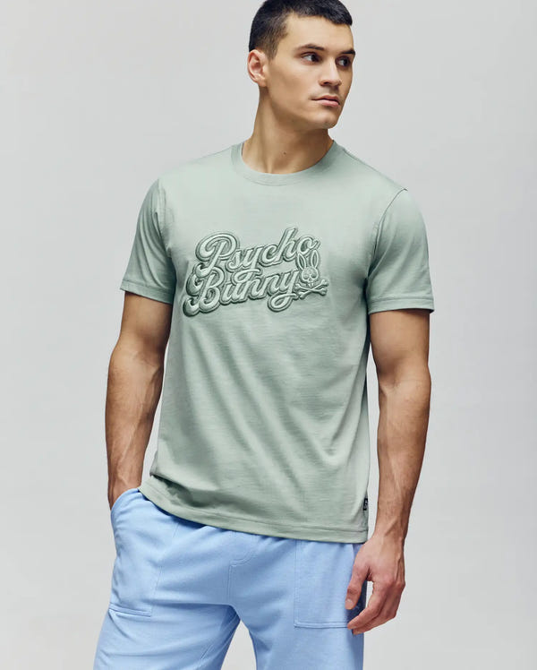 A man in a light green MENS ELIAS GRAPHIC TEE (MT0200047) by Psycho Bunny and light blue pants stands against a plain background, looking left with a neutral expression.