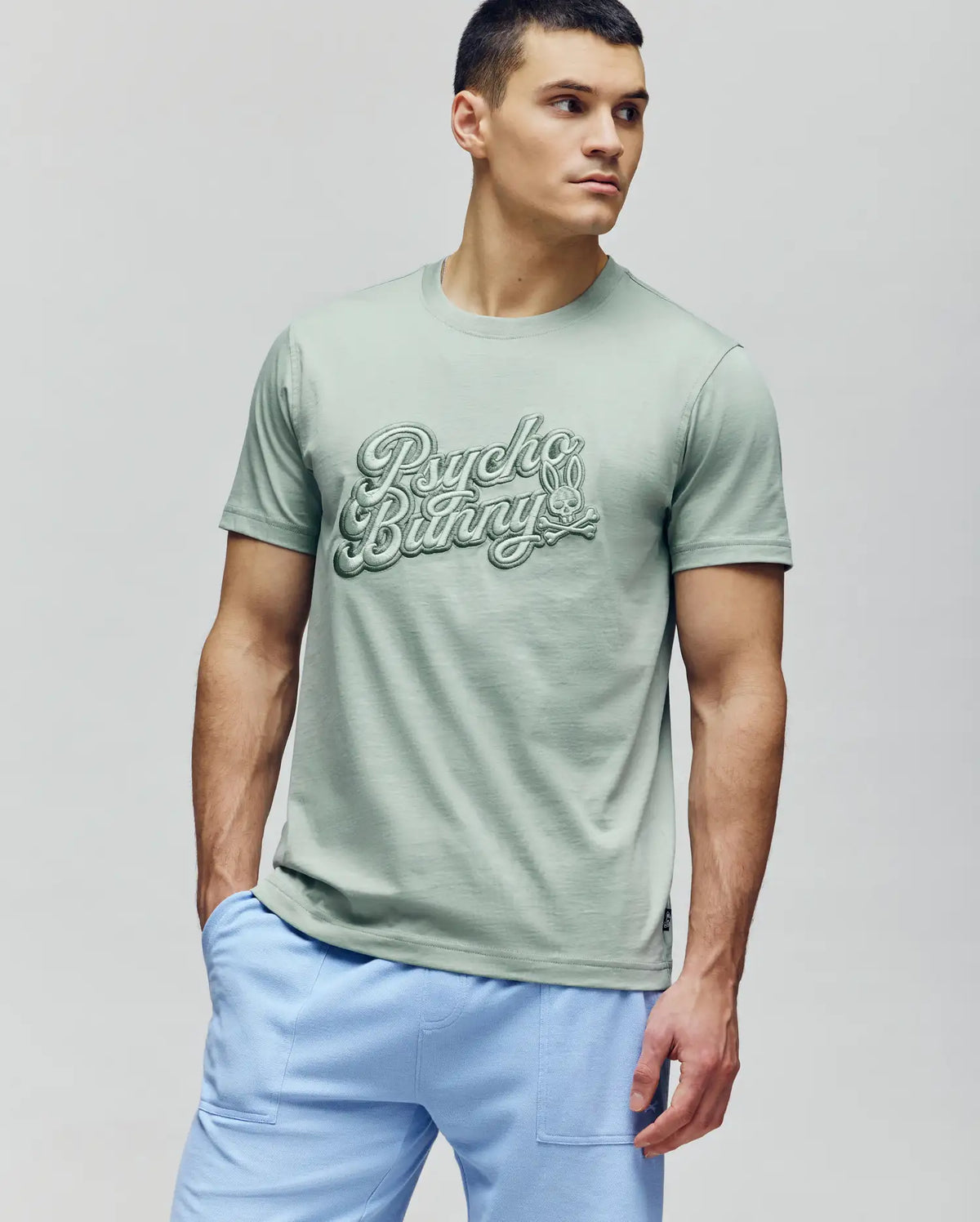 A man in a light green MENS ELIAS GRAPHIC TEE (MT0200047) by Psycho Bunny and light blue pants stands against a plain background, looking left with a neutral expression.