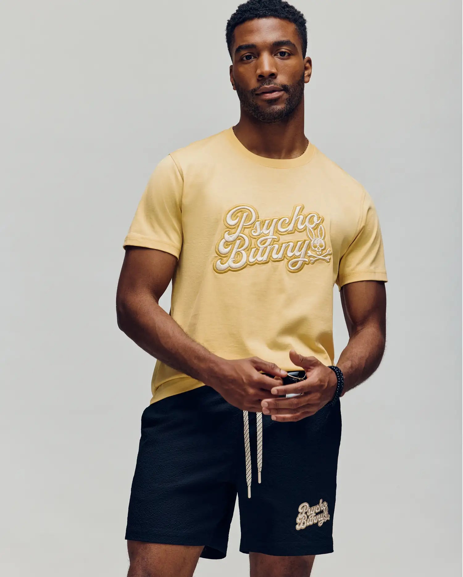 A man with short hair and a trimmed beard stands against a plain background wearing the Psycho Bunny MENS ELIAS GRAPHIC TEE - MT0200047 in yellow Pima cotton and black shorts with matching branding, holding sunglasses in one hand.