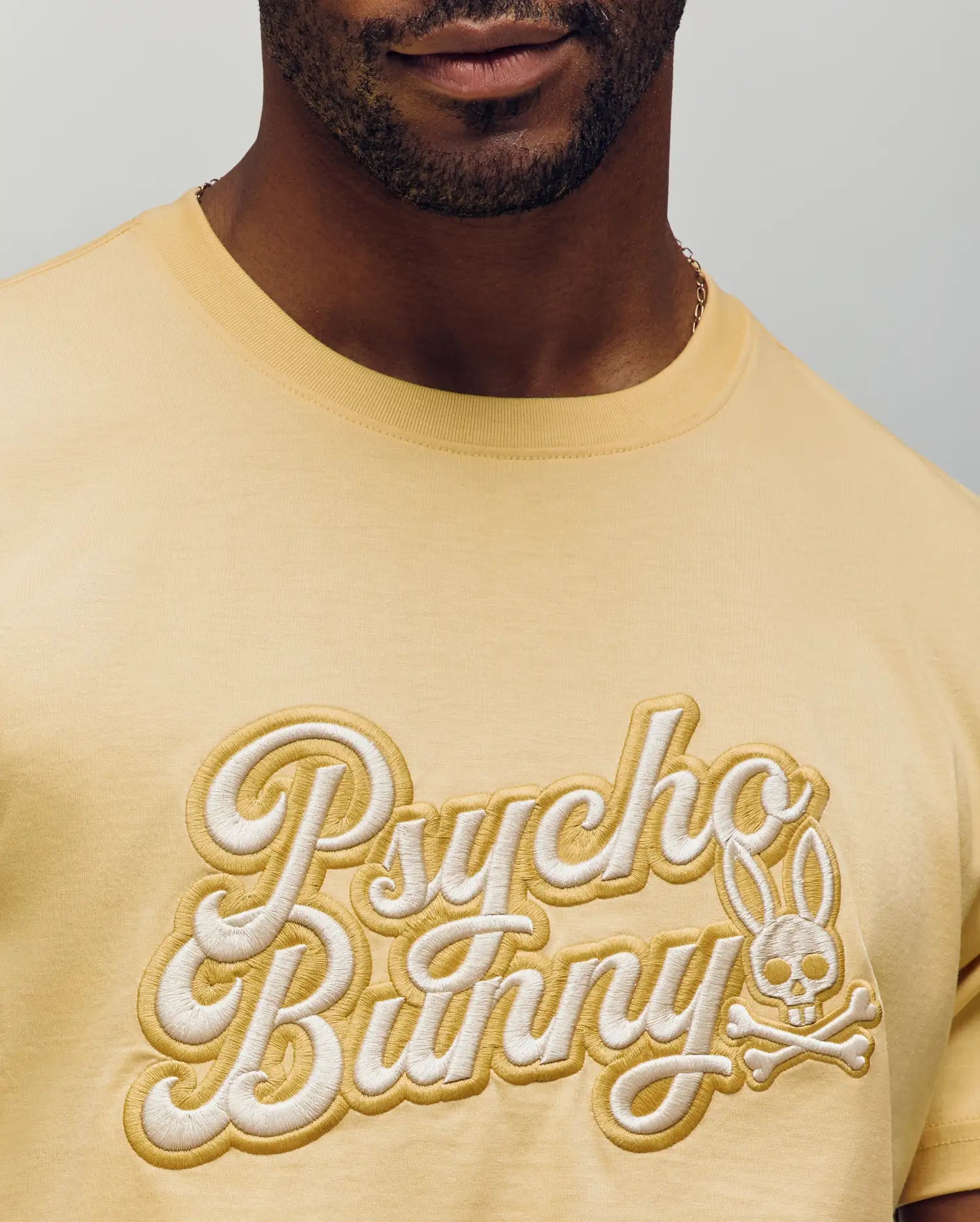 A man wears the Psycho Bunny MENS ELIAS GRAPHIC TEE (MT0200047) in pale yellow, featuring embroidered "Psycho Bunny" text and a bunny skull-and-crossbones graphic on the chest; only his lower face and upper torso are visible.
