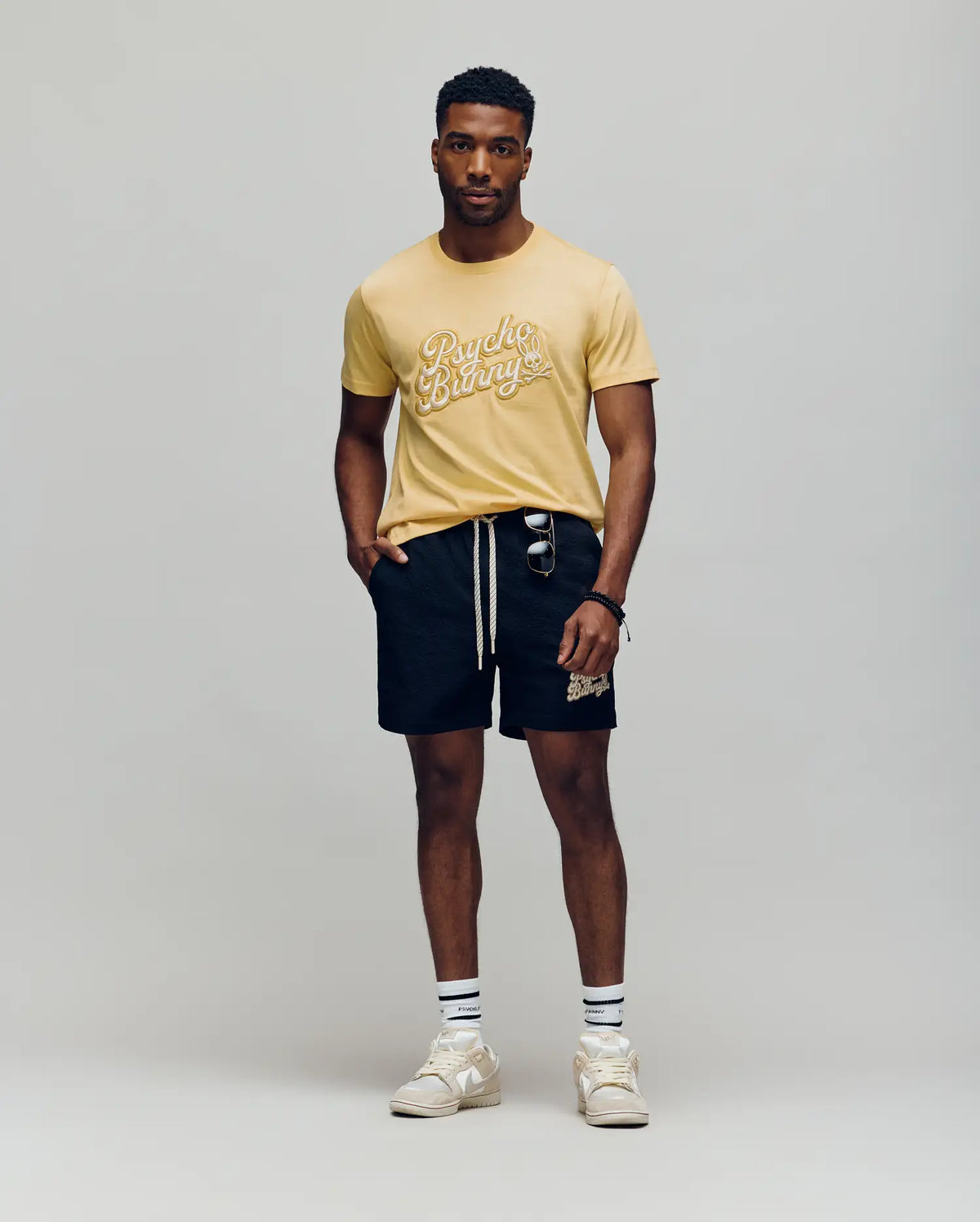 A man stands against a plain background wearing the Psycho Bunny MENS ELIAS GRAPHIC TEE - MT0200047 in yellow with a white Bunny graphic, black drawstring shorts, striped retro crew socks, white sneakers, and sunglasses hanging from his shorts pocket.
