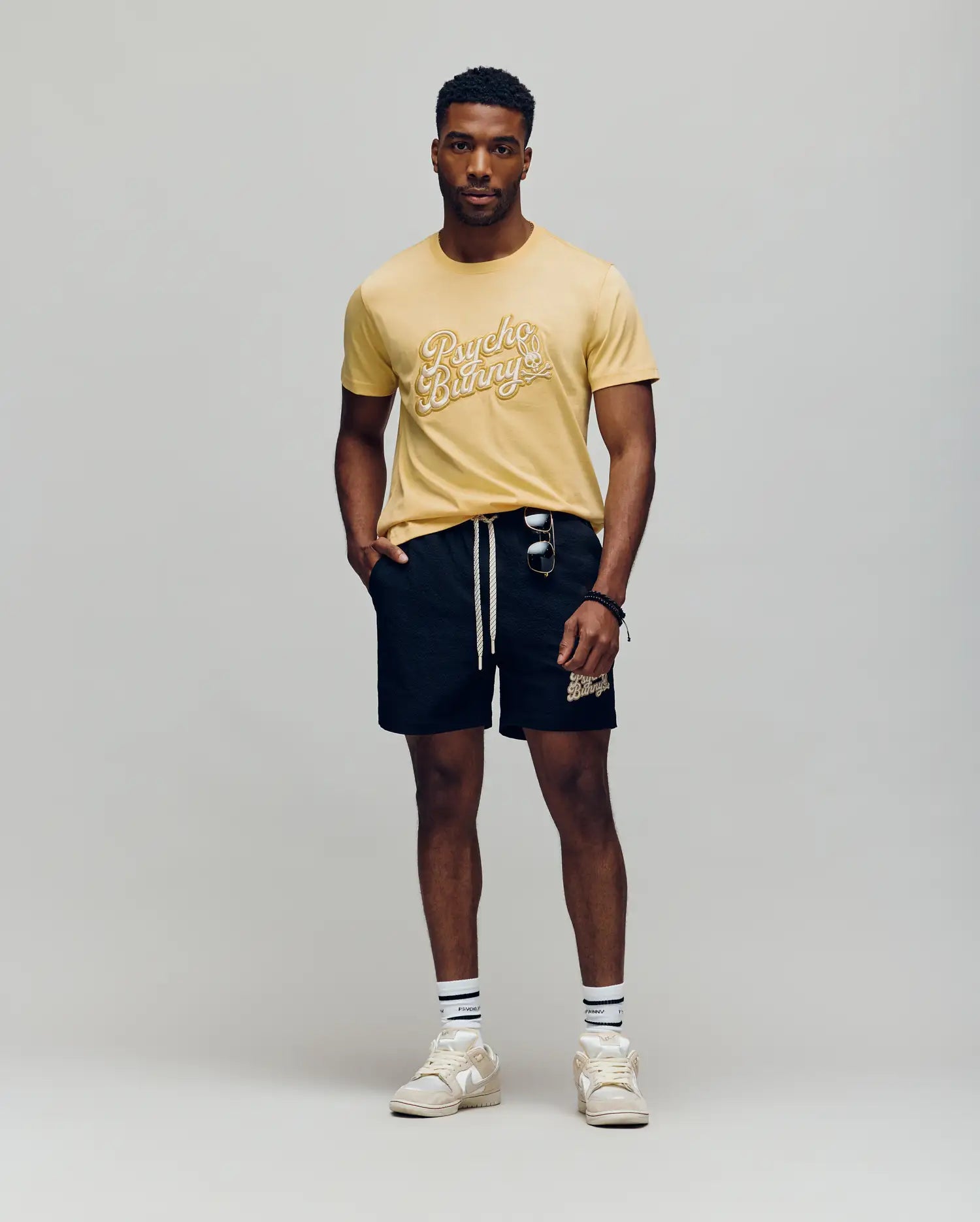 A man stands against a plain background wearing the Psycho Bunny MENS ELIAS GRAPHIC TEE - MT0200047 in yellow with a white Bunny graphic, black drawstring shorts, striped retro crew socks, white sneakers, and sunglasses hanging from his shorts pocket.