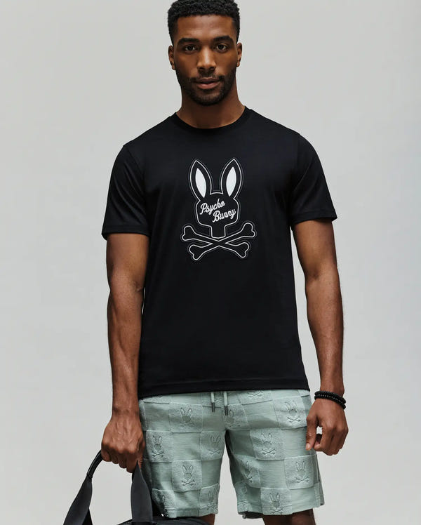 A man wears a Psycho Bunny MENS DEREK CENTER GRAPHIC TEE (MT0200048) in black with an embroidered bunny and crossbones logo, paired with light green bunny-patterned shorts, standing against a plain background holding a black bag.