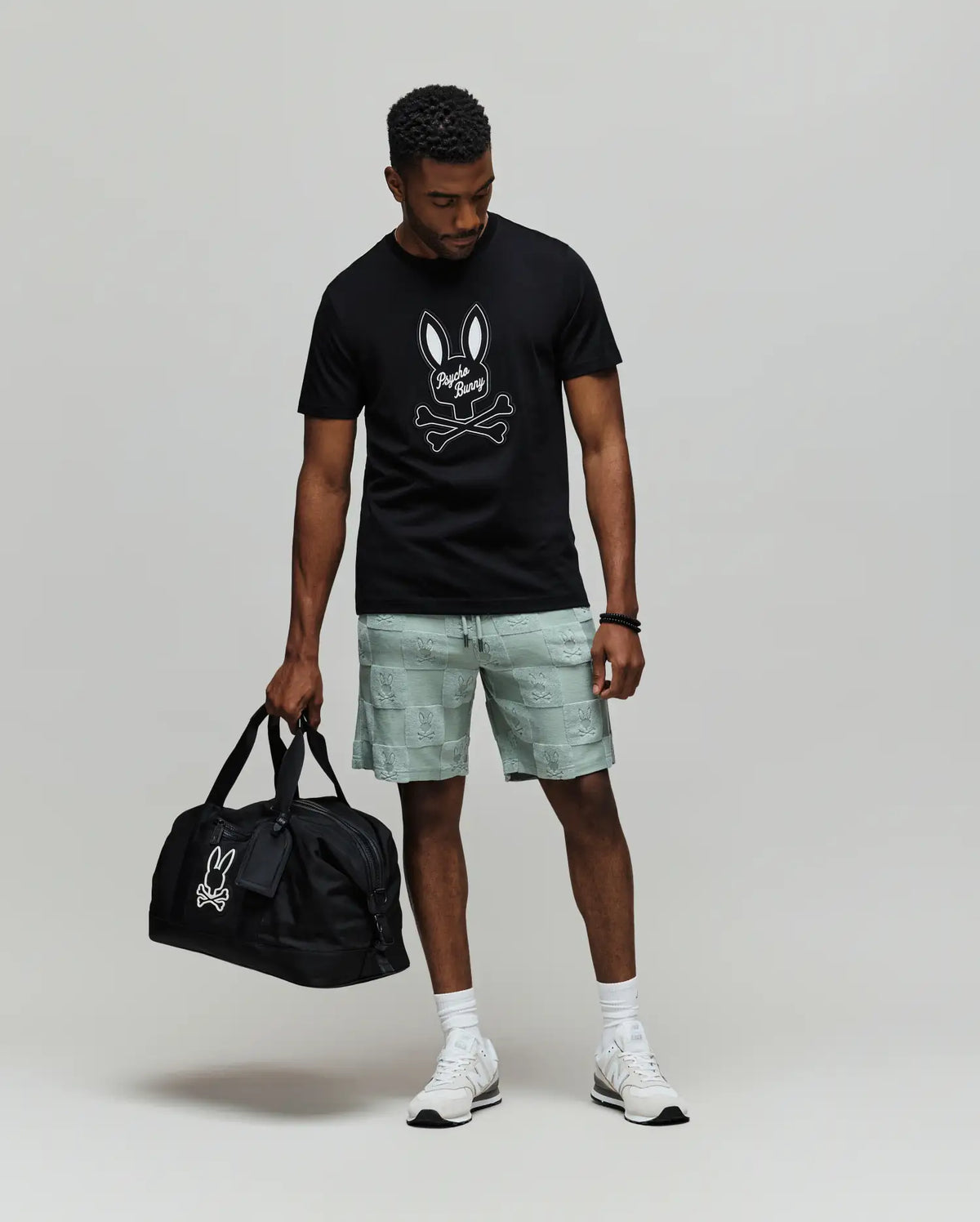 A man wears the Psycho Bunny MENS DEREK CENTER GRAPHIC TEE (MT0200048), featuring a black Pima cotton shirt with an embroidered bunny skull logo, light gray shorts, white socks and sneakers, holding a matching black duffle bag.