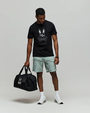 A man wears the Psycho Bunny MENS DEREK CENTER GRAPHIC TEE (MT0200048), featuring a black Pima cotton shirt with an embroidered bunny skull logo, light gray shorts, white socks and sneakers, holding a matching black duffle bag.