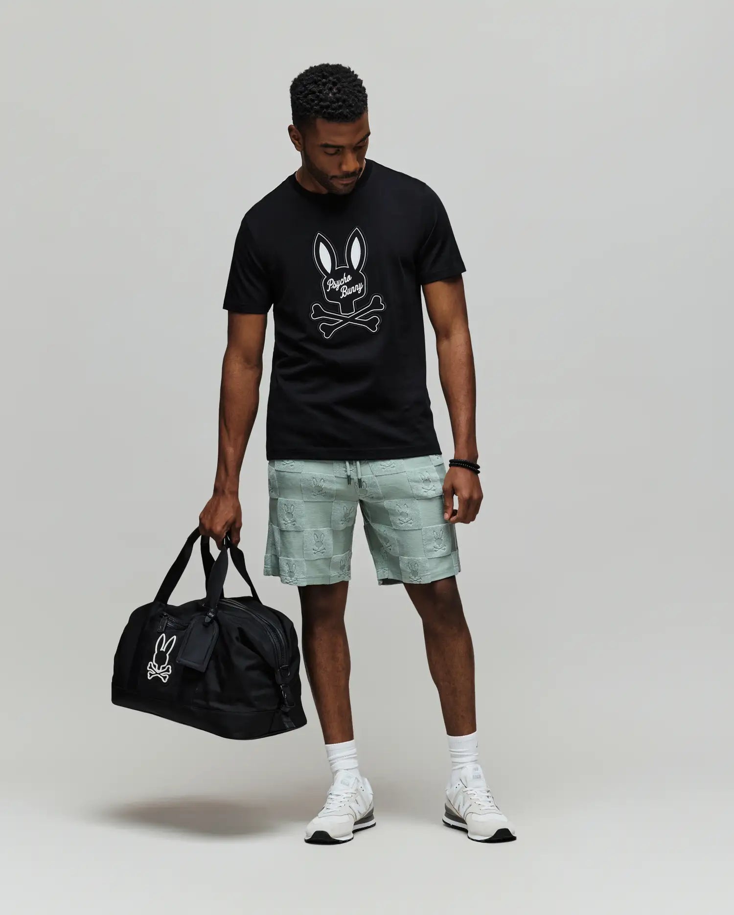 A man wears the Psycho Bunny MENS DEREK CENTER GRAPHIC TEE (MT0200048), featuring a black Pima cotton shirt with an embroidered bunny skull logo, light gray shorts, white socks and sneakers, holding a matching black duffle bag.