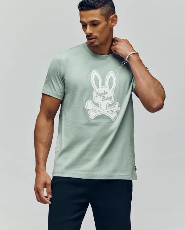 A man in the Psycho Bunny MENS DEREK CENTER GRAPHIC TEE - MT0200048, a light green Pima cotton shirt with a large white Bunny skull and crossbones and "Psycho Bunny" text, pairs it with black pants against a plain light background.