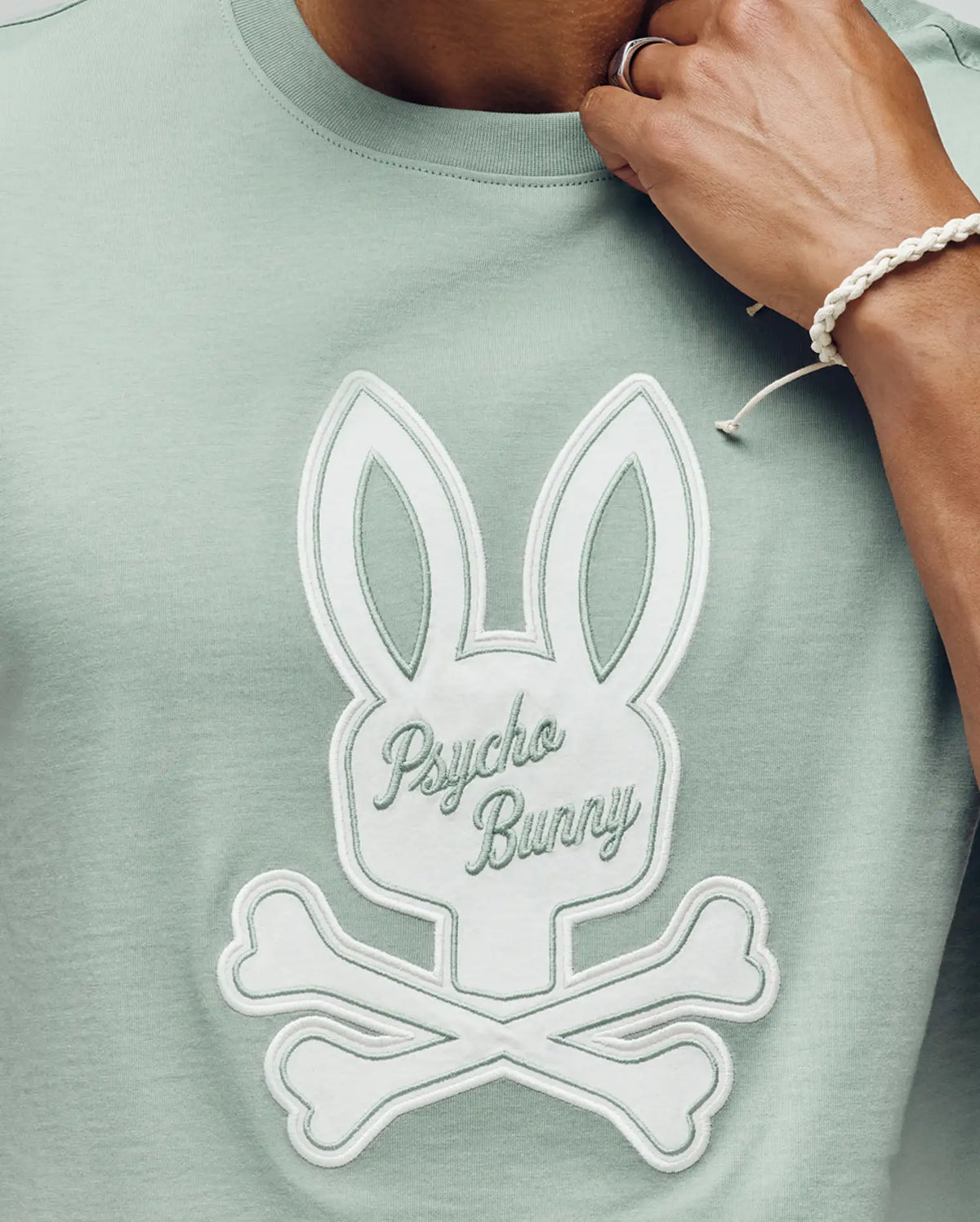 A person wears the Psycho Bunny MENS DEREK CENTER GRAPHIC TEE (MT0200048), a light green Pima cotton T-shirt with a large white embroidered bunny skull and crossbones logo and "Psycho Bunny" on the chest. Their ring-adorned hand is near their face.
