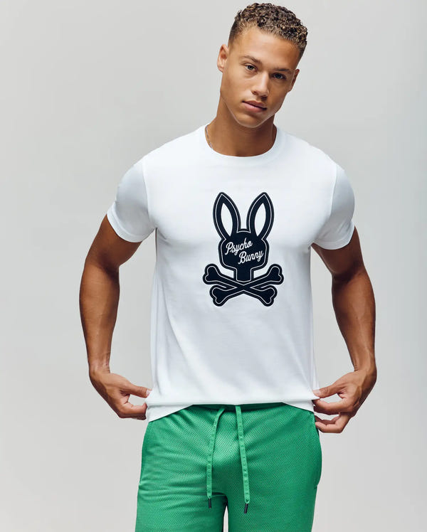 A young man stands against a plain light background, wearing Psycho Bunny's MENS DEREK CENTER GRAPHIC TEE (MT0200048)—a white Pima cotton T-shirt with a bold black bunny skull and crossbones graphic—paired with green drawstring shorts.