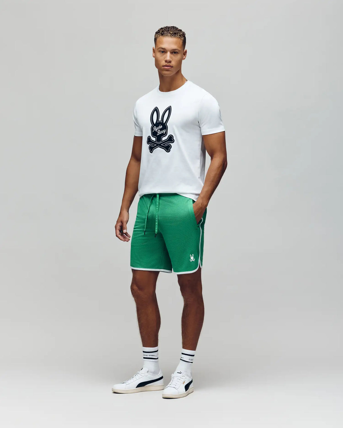 A man stands facing forward in a Psycho Bunny MENS DEREK CENTER GRAPHIC TEE - MT0200048, green athletic shorts with white trim, white socks with dark stripes, and white sneakers against a plain gray background.