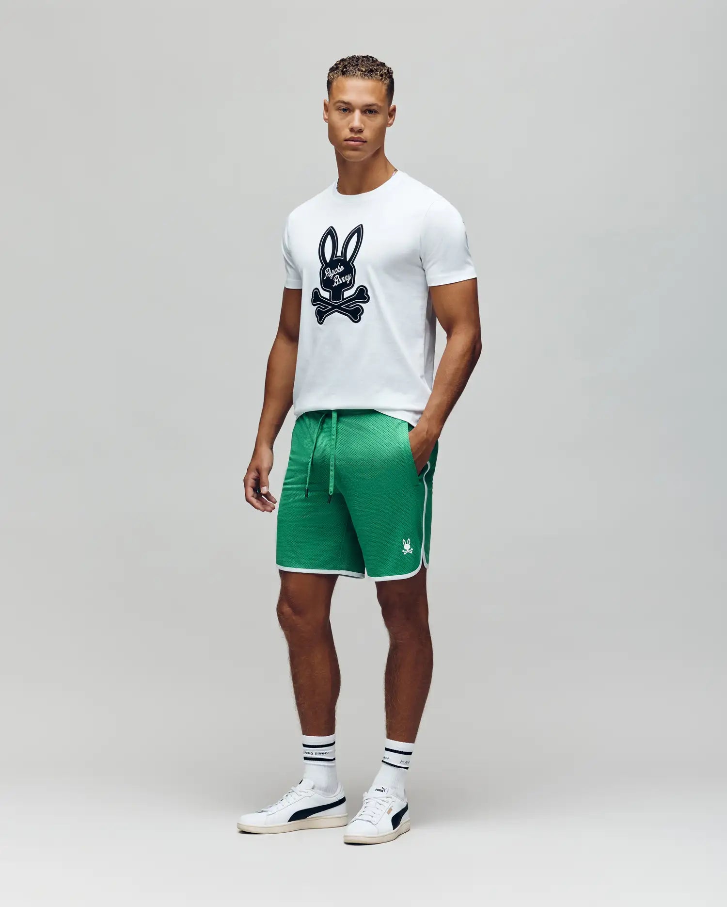 A man stands facing forward in a Psycho Bunny MENS DEREK CENTER GRAPHIC TEE - MT0200048, green athletic shorts with white trim, white socks with dark stripes, and white sneakers against a plain gray background.