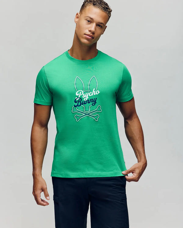 A man wears the Psycho Bunny MENS GARRET GRAPHIC TEE (MT0200050) featuring a bunny ears and crossbones graphic against a plain light background. He pairs this green Pima cotton tee with dark shorts.