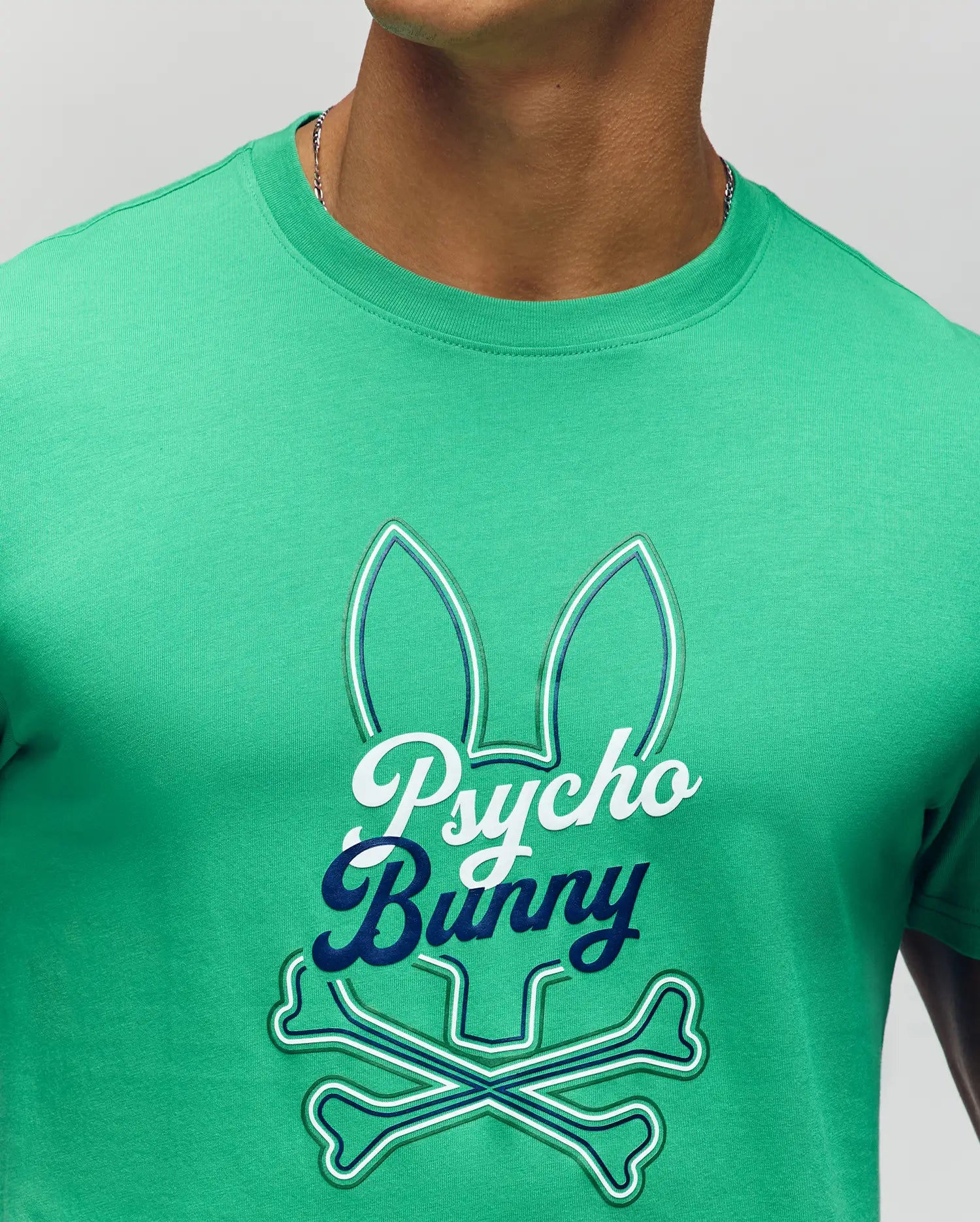 A person wears the Psycho Bunny MENS GARRET GRAPHIC TEE (MT0200050), a green crewneck made from soft Pima cotton, featuring a bunny ears outline with crossbones and bold "Psycho Bunny" script on the front.
