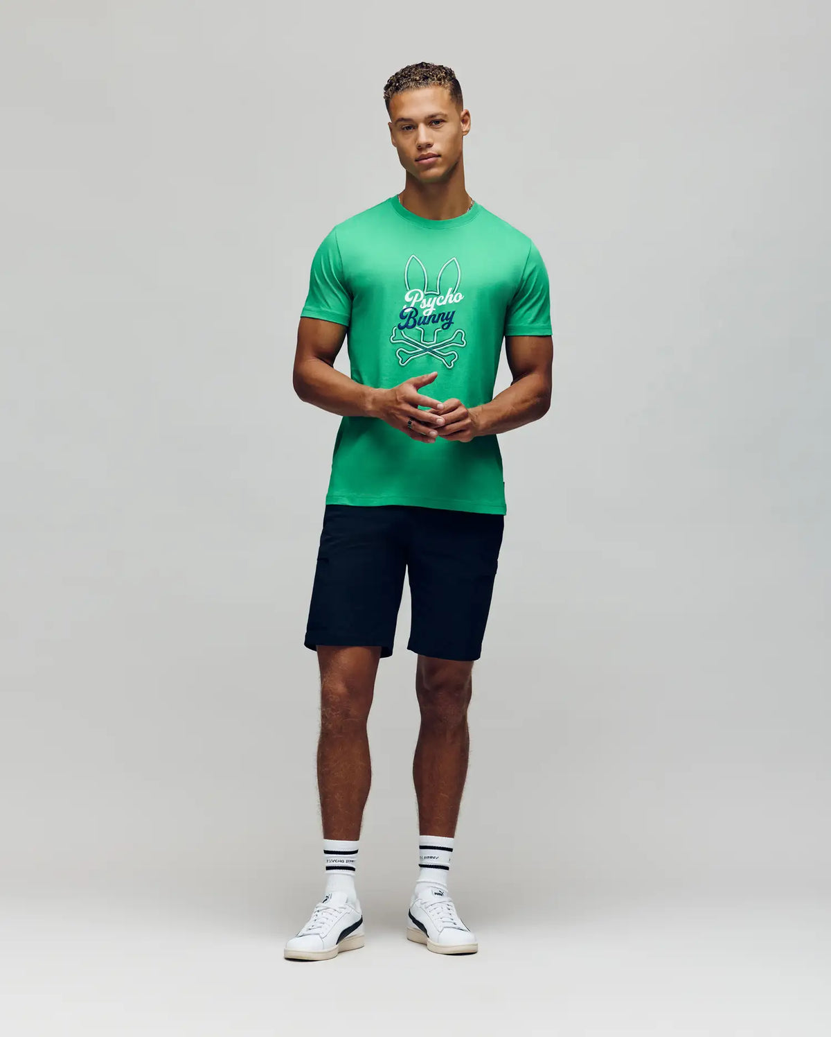 A man faces forward against a light gray background, wearing a bright green Psycho Bunny MENS GARRET GRAPHIC TEE (MT0200050), dark shorts, white crew socks, and white sneakers.