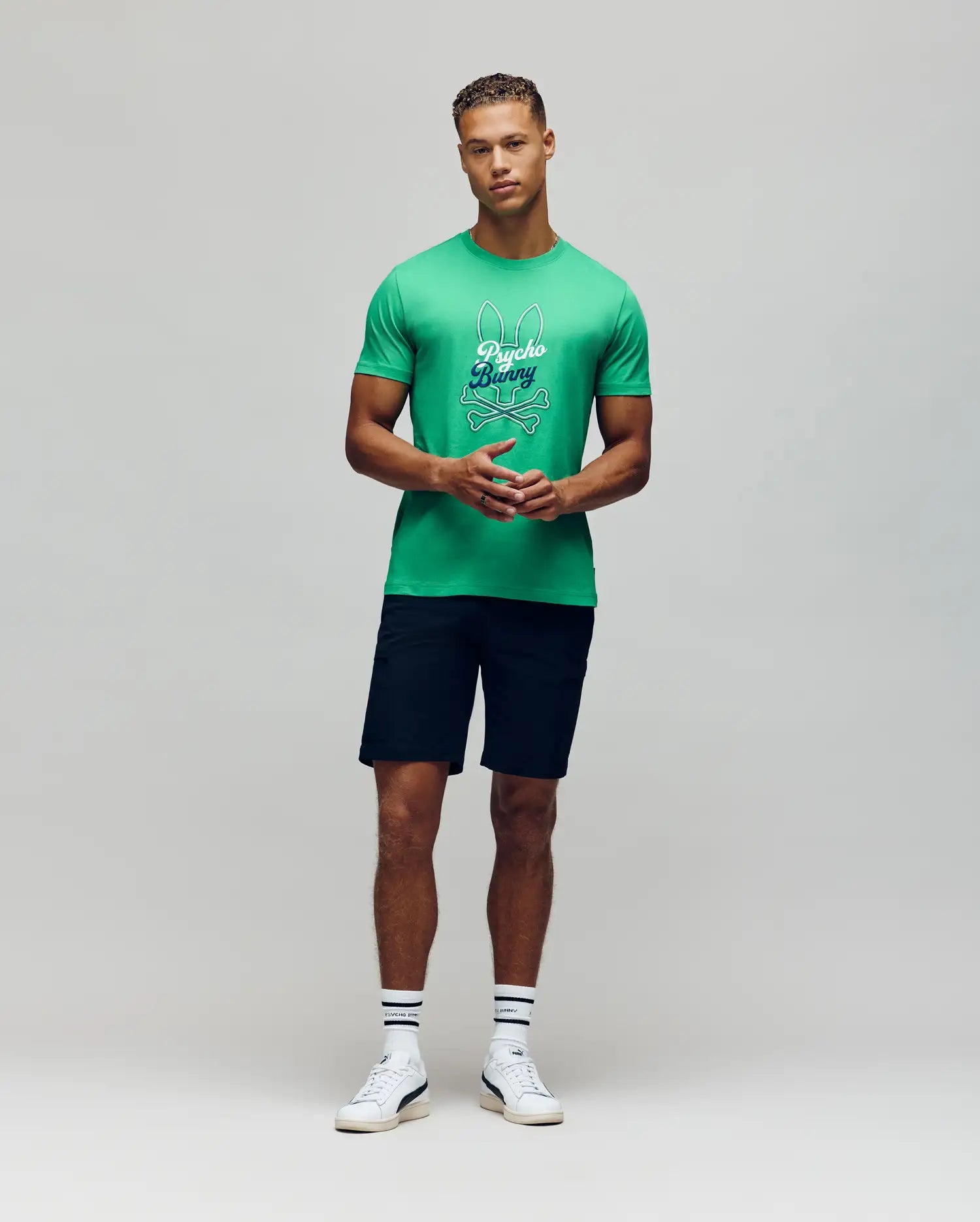 A man faces forward against a light gray background, wearing a bright green Psycho Bunny MENS GARRET GRAPHIC TEE (MT0200050), dark shorts, white crew socks, and white sneakers.