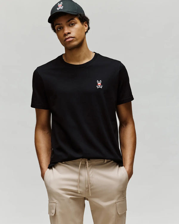 A person wearing a black cap, Psycho Bunny MENS MISCHIEF BUNNY TEE (MT0200051) featuring an embroidered bunny logo on the chest, and beige cargo pants stands against a plain light gray background.