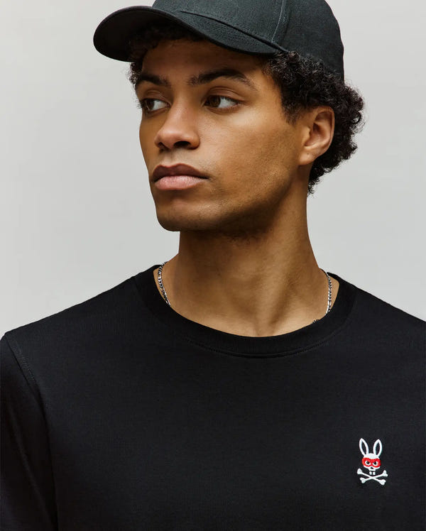 A young man wears the Psycho Bunny MENS MISCHIEF BUNNY TEE (MT0200051) in black, made from Pima cotton, featuring a small embroidered white Mischief Bunny with red eyes and crossbones on the chest, as he looks to his left.