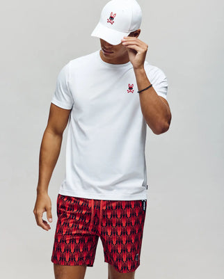 A man wears the Psycho Bunny MENS MISCHIEF BUNNY TEE (MT0200051) in white, featuring an embroidered bunny logo, with red and black shorts and a white cap with a red bunny logo, standing against a plain background holding his cap.