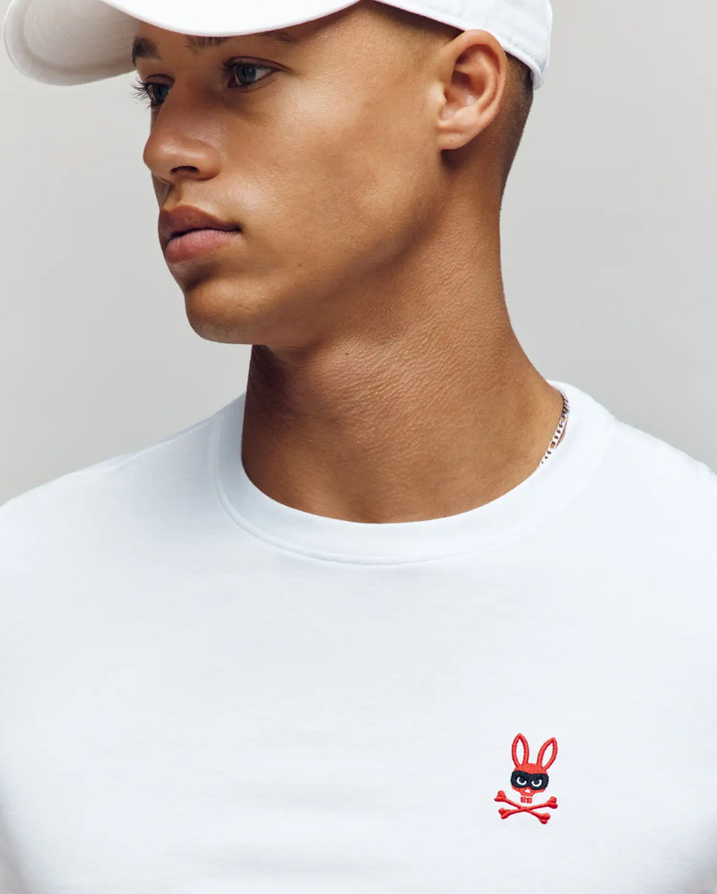 A man in a white cap and Psycho Bunny MENS MISCHIEF BUNNY TEE (MT0200051) with an embroidered bunny and crossbones logo on the chest, looks to the side against a neutral background.
