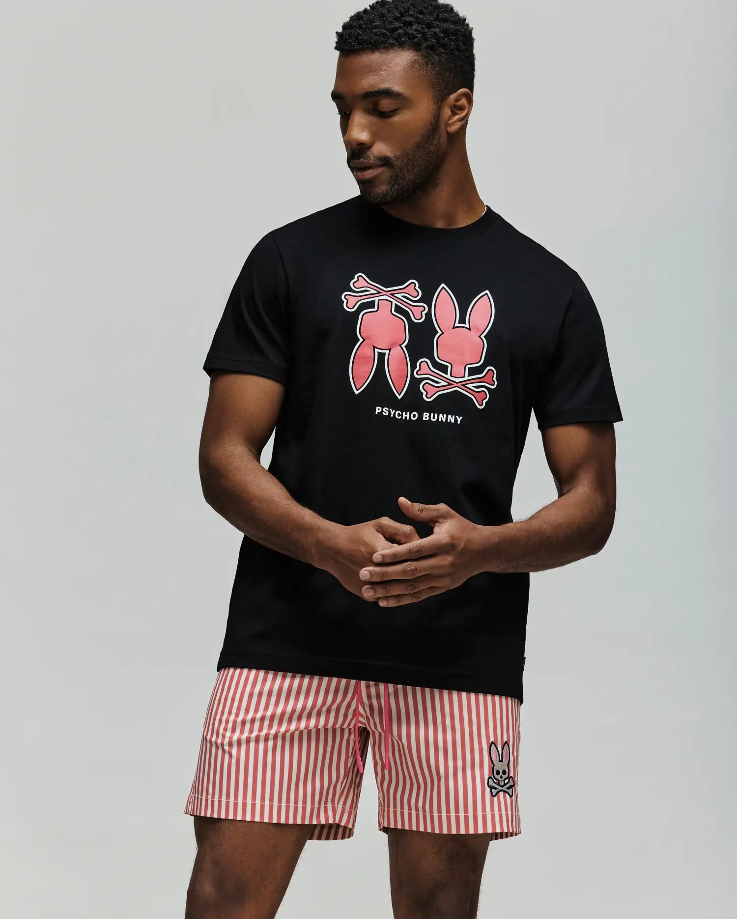 A man stands with his hands by his sides, wearing the Psycho Bunny MENS DAWSON GRAPHIC TEE (MT0200053) in black Pima cotton with large pink bunny graphics and text, paired with pink and white striped shorts featuring a small Bunny logo.