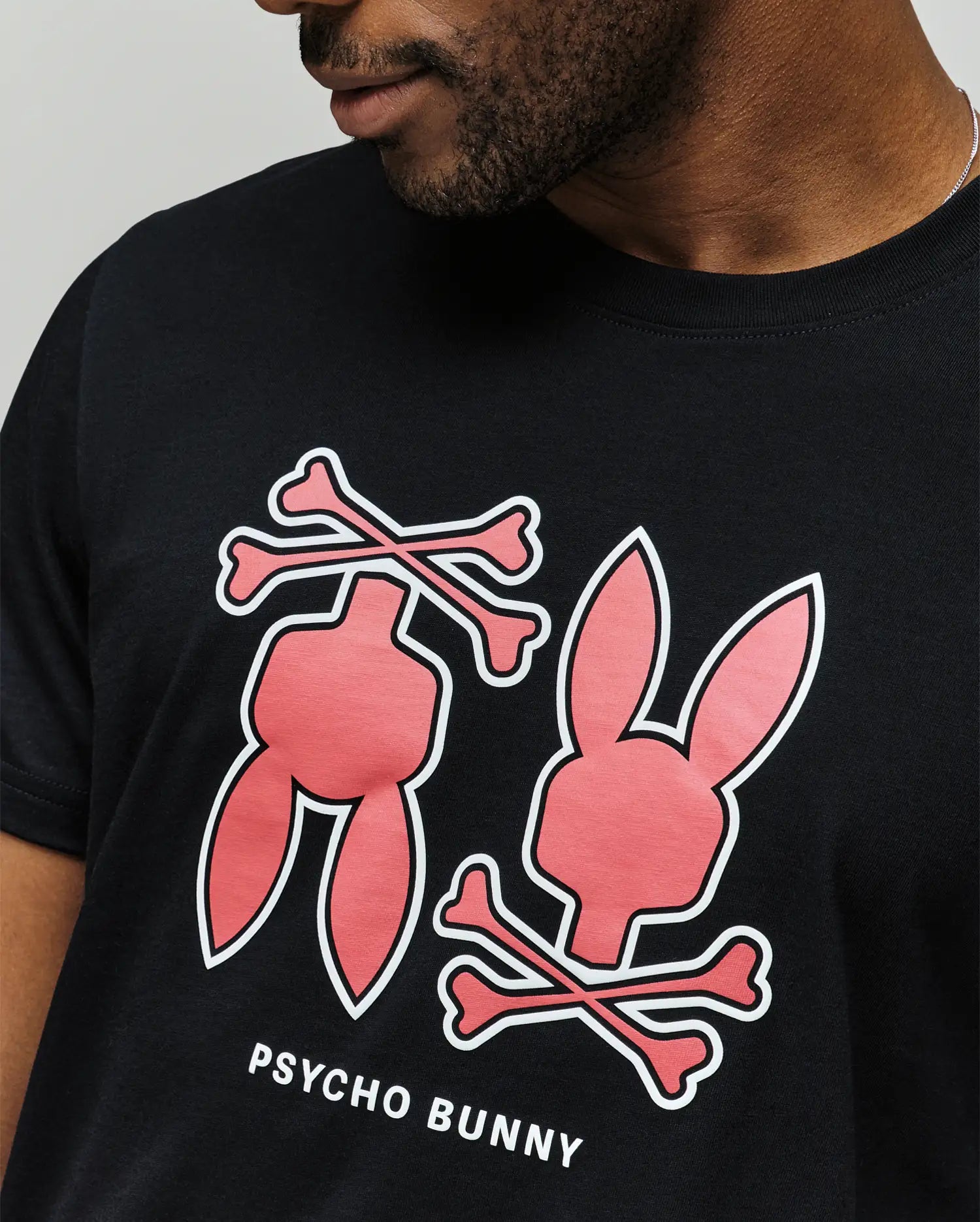 Wearing the Psycho Bunny MENS DAWSON GRAPHIC TEE (MT0200053), a soft black Pima cotton shirt featuring the iconic logo and two pink bunny graphics above white crossbones—one with just the head and another with only the body.