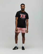 A man stands in a studio wearing the Psycho Bunny MENS DAWSON GRAPHIC TEE - MT0200053 in black, featuring red Bunny graphics, paired with red and white striped shorts, white socks, and black Adidas sneakers against a plain light gray background.