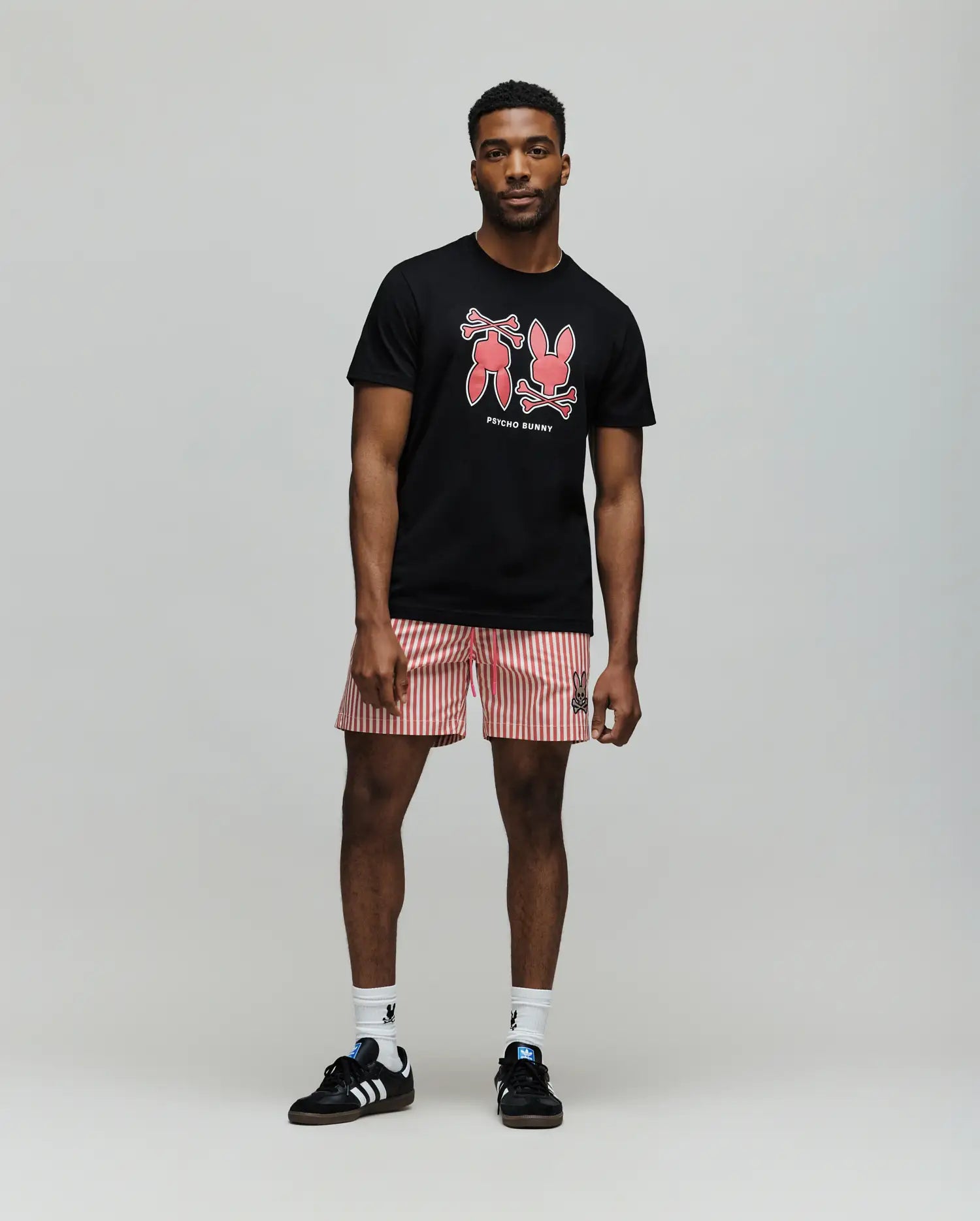 A man stands in a studio wearing the Psycho Bunny MENS DAWSON GRAPHIC TEE - MT0200053 in black, featuring red Bunny graphics, paired with red and white striped shorts, white socks, and black Adidas sneakers against a plain light gray background.