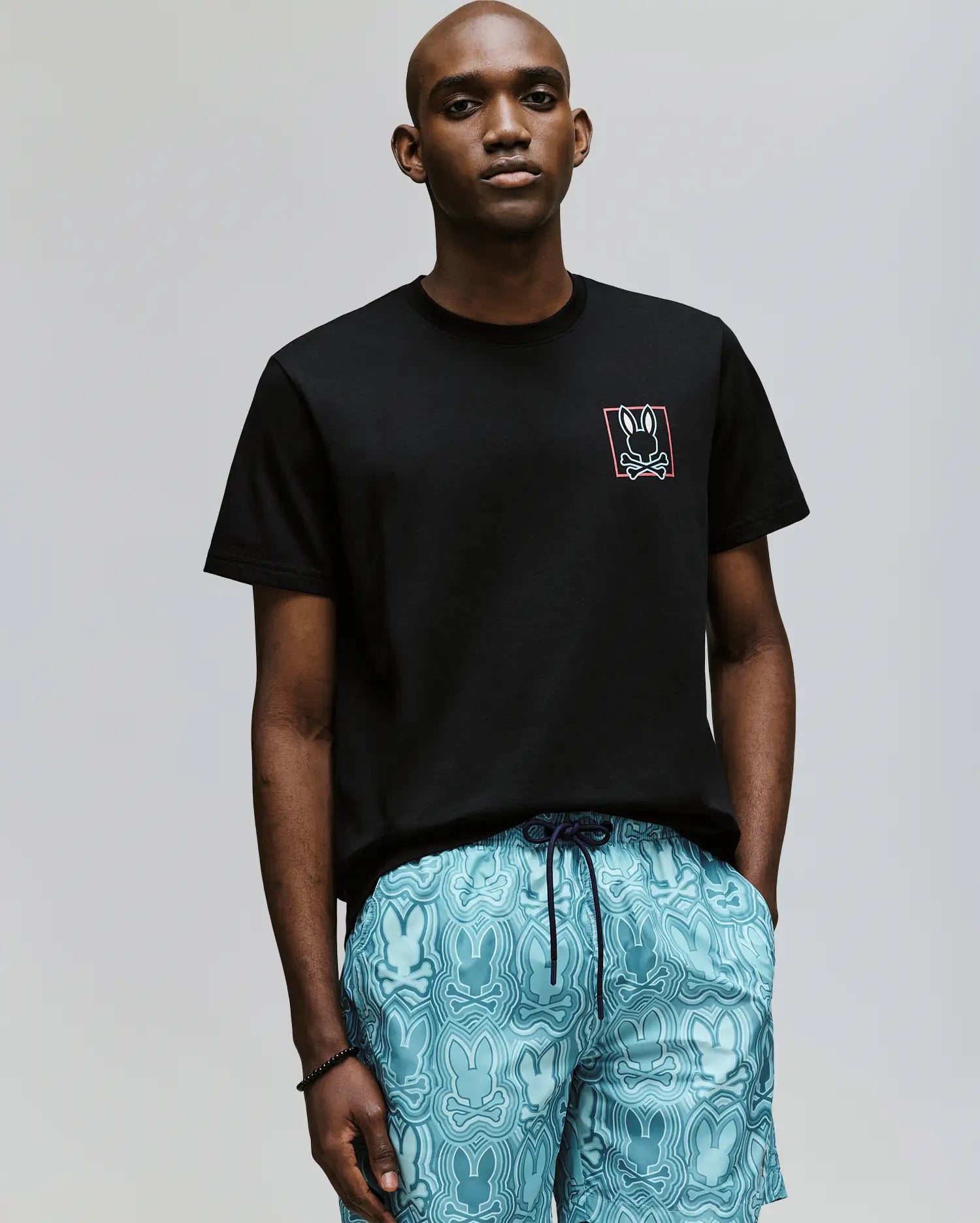 A man stands against a plain background wearing the Psycho Bunny MENS JESSE GRAPHIC TEE (MT0200056) made from eco-friendly Pima cotton, featuring a small white bunny logo, and light blue shorts with a repeating bunny pattern.