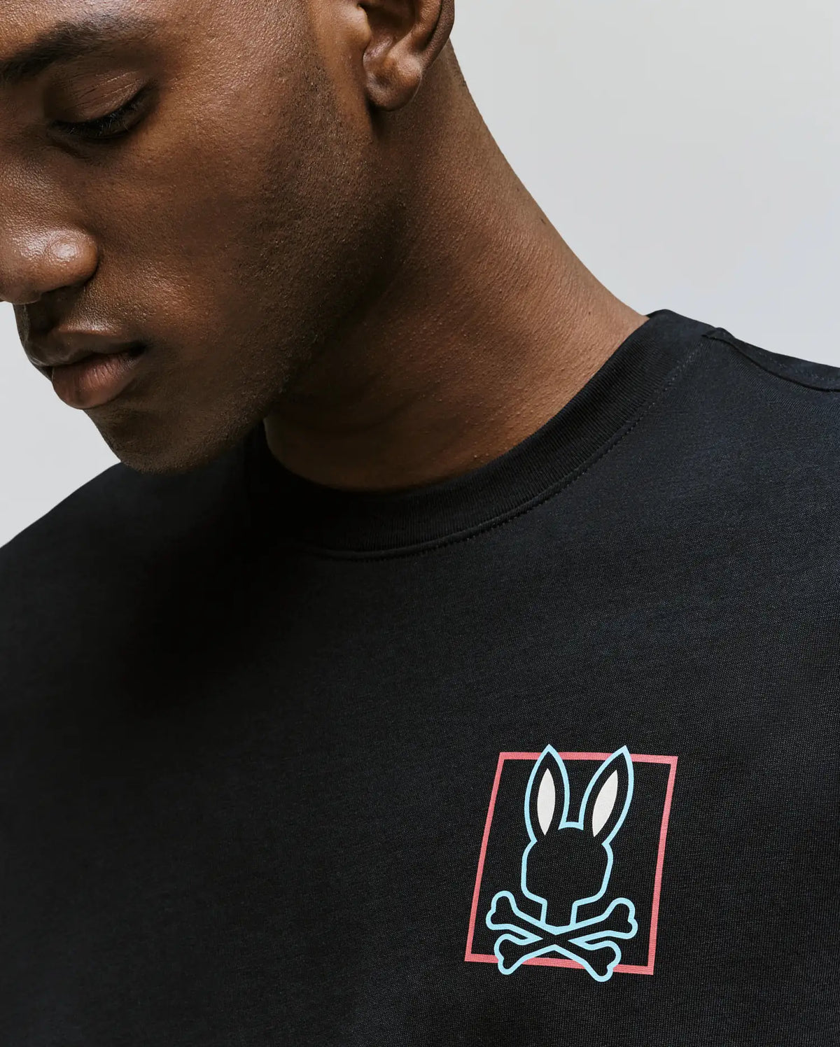 A man wearing the Psycho Bunny MENS JESSE GRAPHIC TEE (MT0200056), an eco-friendly Pima cotton shirt with a bunny head above crossed bones, printed with water-based inks inside a red-bordered rectangle on the chest.