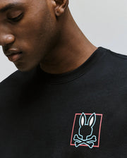 A man wearing the Psycho Bunny MENS JESSE GRAPHIC TEE (MT0200056), an eco-friendly Pima cotton shirt with a bunny head above crossed bones, printed with water-based inks inside a red-bordered rectangle on the chest.