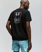 A man faces away wearing the Psycho Bunny MEN'S JESSE GRAPHIC TEE (MT0200056), a black Pima cotton shirt with a large bunny skull and “Psycho Bunny Since 2003” in eco-friendly water-based print, paired with turquoise bunny-patterned shorts.