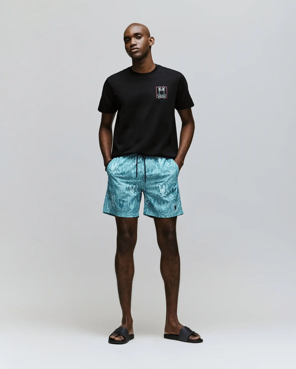 A man faces forward against a plain background, wearing the Psycho Bunny MENS JESSE GRAPHIC TEE (MT0200056) in black with water-based chest prints, turquoise patterned shorts, and black slide sandals. His hands are in his pockets.