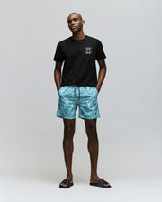 A man faces forward against a plain background, wearing the Psycho Bunny MENS JESSE GRAPHIC TEE (MT0200056) in black with water-based chest prints, turquoise patterned shorts, and black slide sandals. His hands are in his pockets.