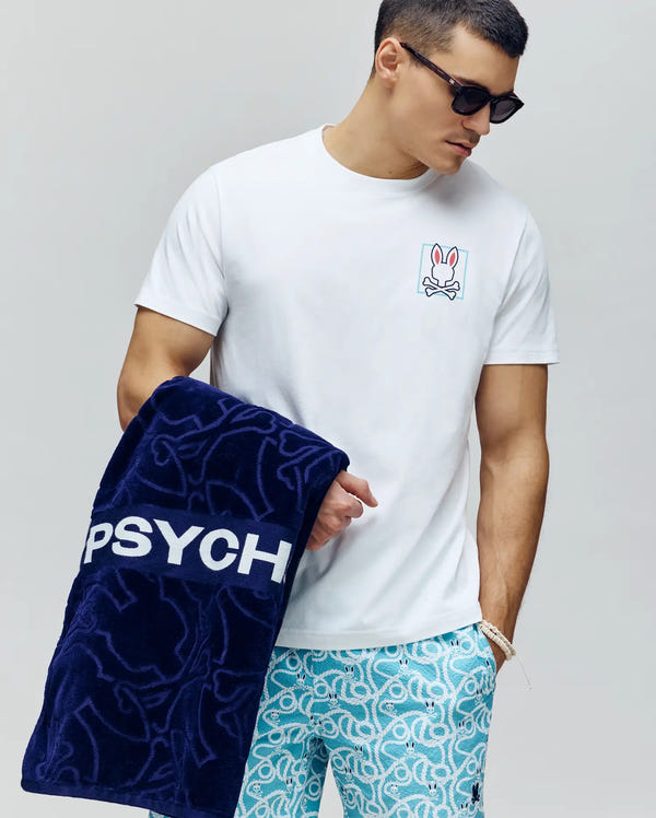 A man wearing sunglasses, a Psycho Bunny MENS JESSE GRAPHIC TEE (MT0200056) with a small bunny graphic and blue patterned shorts holds a large navy towel featuring "PSYCH" in water-based prints.