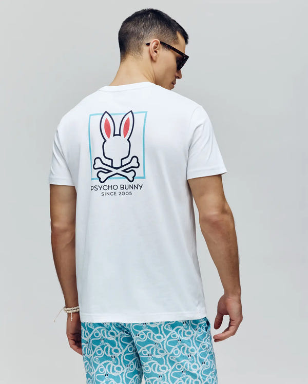 A man in sunglasses and a Psycho Bunny MENS JESSE GRAPHIC TEE (MT0200056), featuring a large bunny skull logo and "Psycho Bunny Since 2005" on the back, with light blue patterned shorts, stands facing away from the camera.