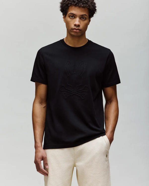 A man with short curly hair wears the Psycho Bunny MENS REMY GRAPHIC TEE (MT0200059) in black, made of soft Pima cotton with an embossed Bunny and crossbones logo, paired with light-colored pants against a light gray background.