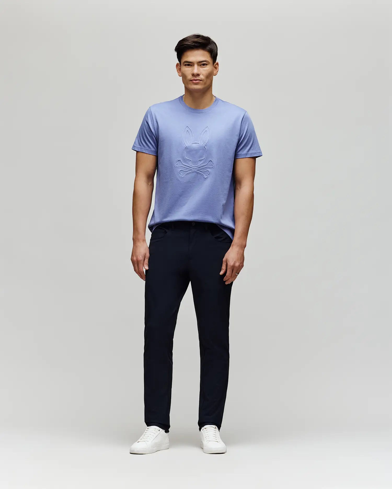 A man stands against a light gray background, wearing the Psycho Bunny MENS REMY GRAPHIC TEE - MT0200059 in light purple Pima cotton, featuring an embossed design and subtle Bunny logo, paired with dark pants and white sneakers. He faces forward neutrally.