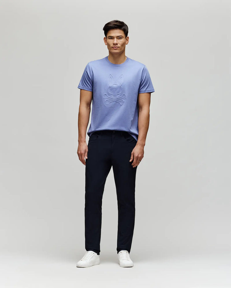 A man stands against a light gray background, wearing the Psycho Bunny MENS REMY GRAPHIC TEE - MT0200059 in light purple Pima cotton, featuring an embossed design and subtle Bunny logo, paired with dark pants and white sneakers. He faces forward neutrally.