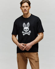 A man with short brown hair stands against a light gray background, wearing tan cargo pants and the Psycho Bunny MENS RYDER OVERSIZED FIT TEE (MT0200062) in black, featuring premium Pima cotton and an embroidered Bunny skull graphic.